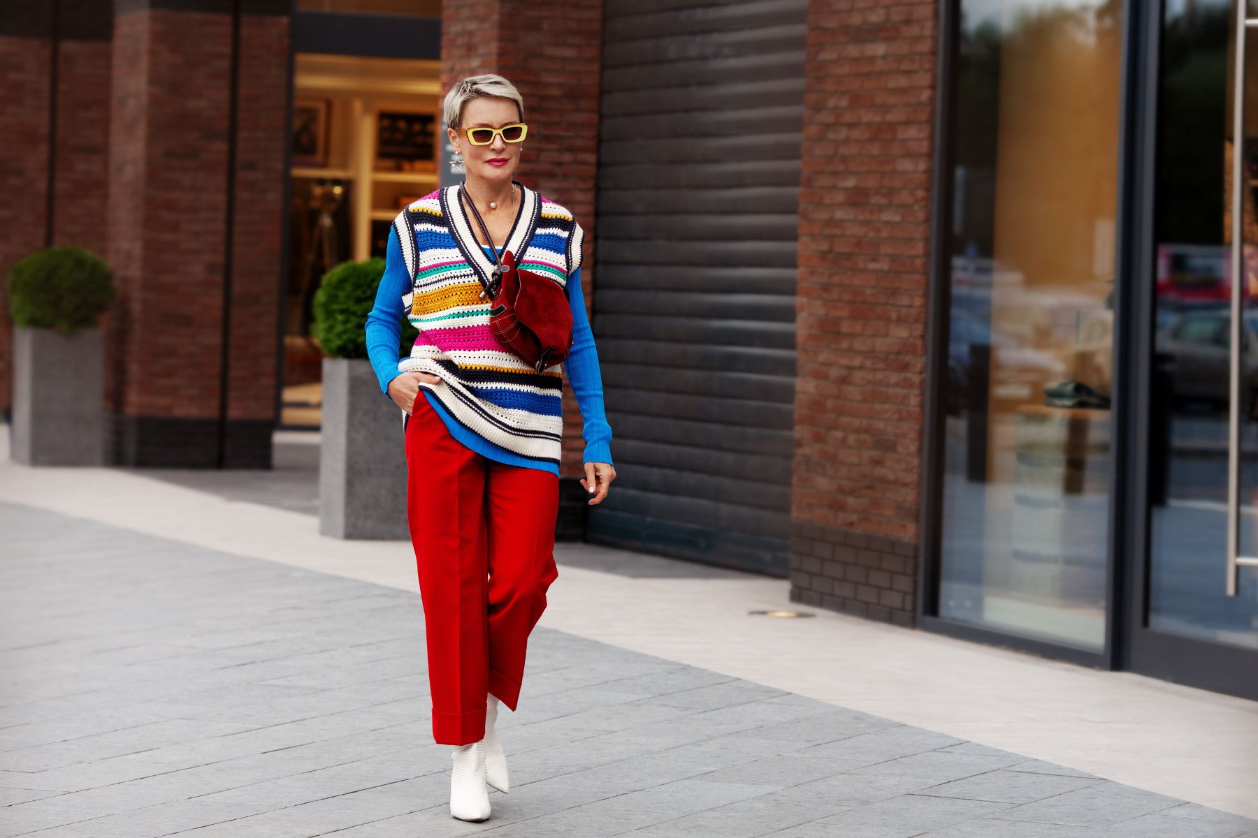 Fashionable Woman wearing trendy outfit, red trousers, blue jacket, striped vest, white shoes and yellow sunglasses. Model with short hair walking the street